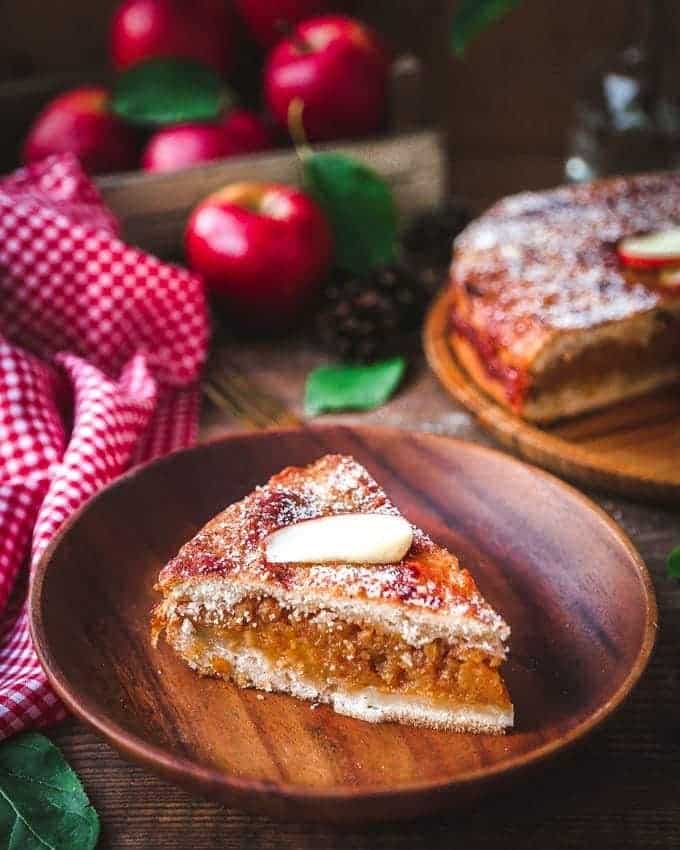 A slice of pie on a wooden plates with apples in the background.