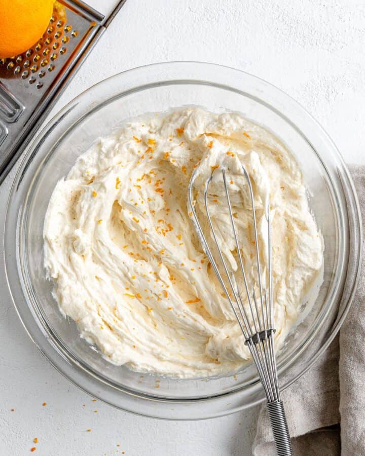A bowl of fluffy white frosting with orange zest on top and a whisk.