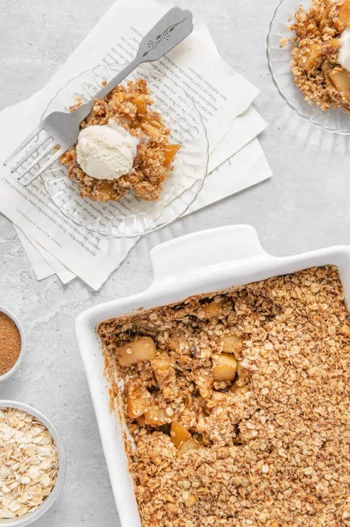 Two portions of apple crisp on plates next to the remaining crisp in a baking dish.