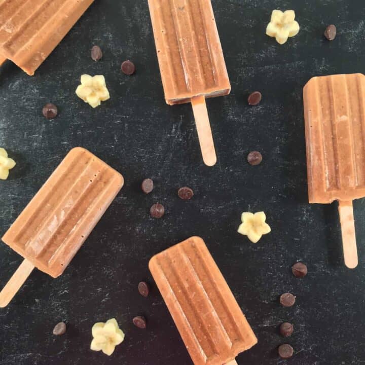 Popsicles on a dark surface next to chocolate chips and banana pieces.