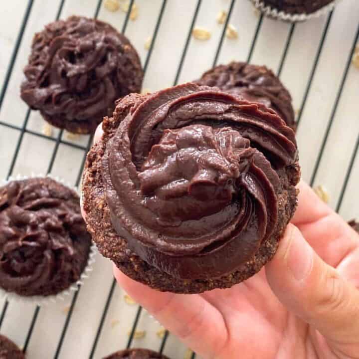 A hand holds up a chocolate cupcake decorated with frosting.