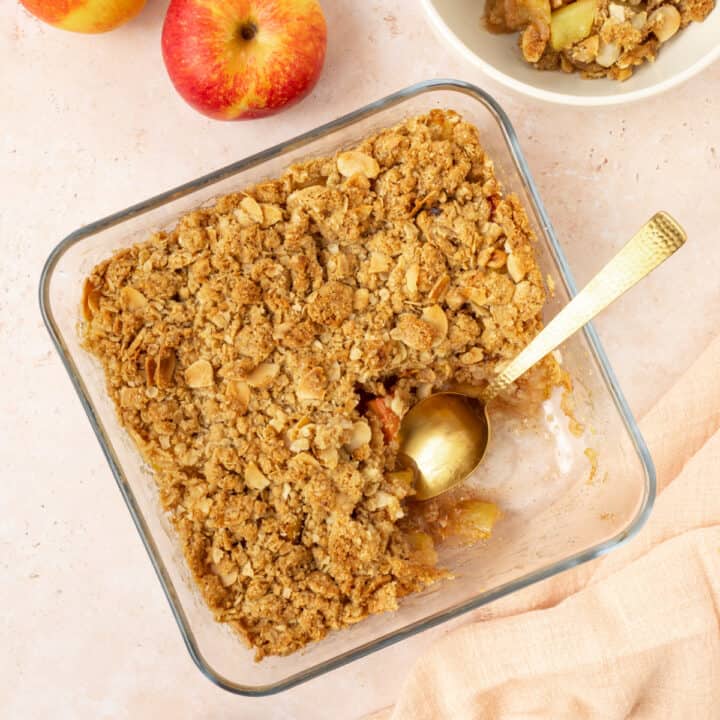 Vegan apple crisp in a dish next to apples, a pink napkin and a spoon.
