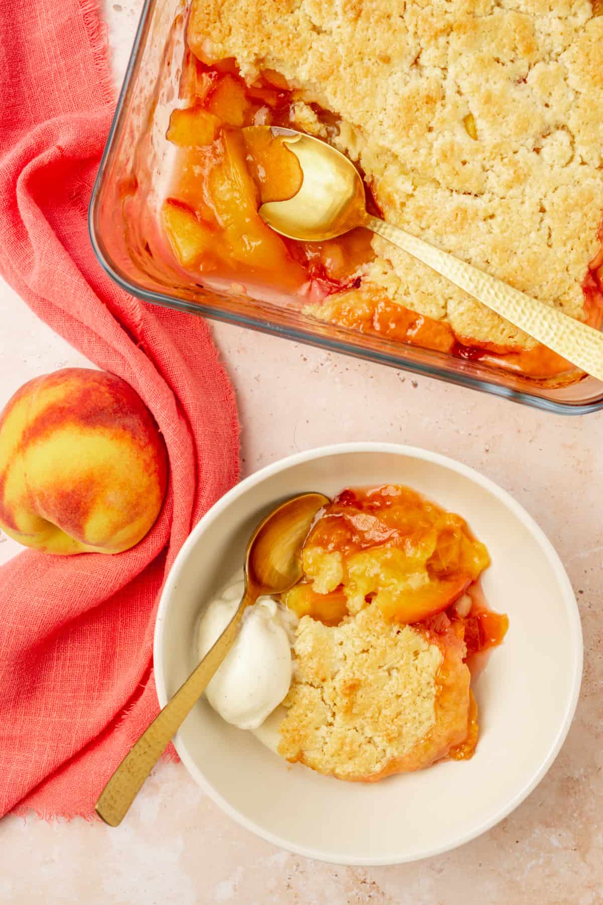 A dish of peach cobbler, next to a serving in a bowl and a pink napkin.