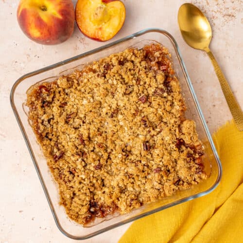 A peach crisp in a square dish next to peaches, a yellow napkin and a spoon.