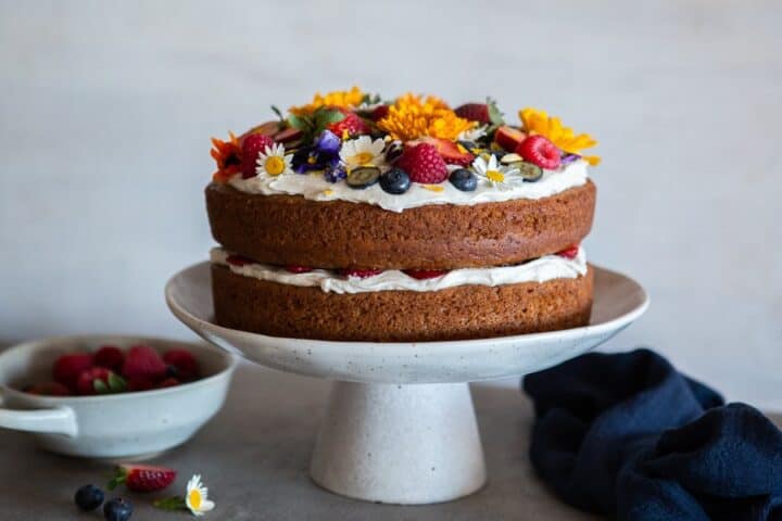 A two layer vanilla cake with coconut cream, berries and edible flowers on a cake stand.