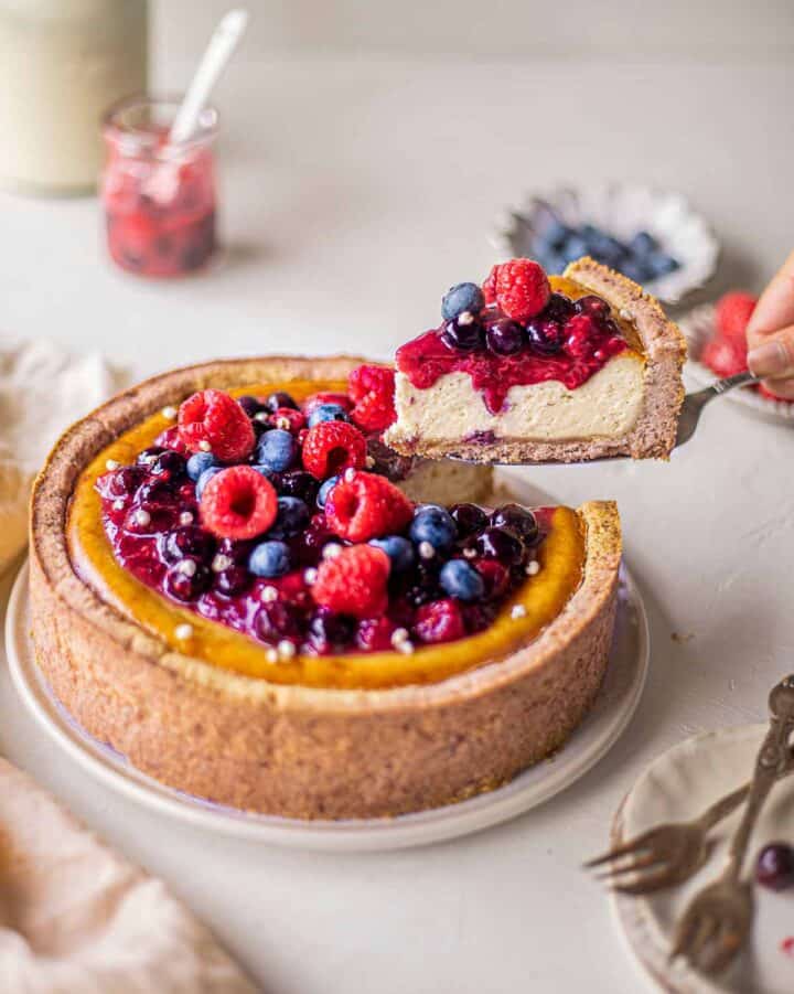 A hand holds up a slice of baked cheesecake topped with berries.