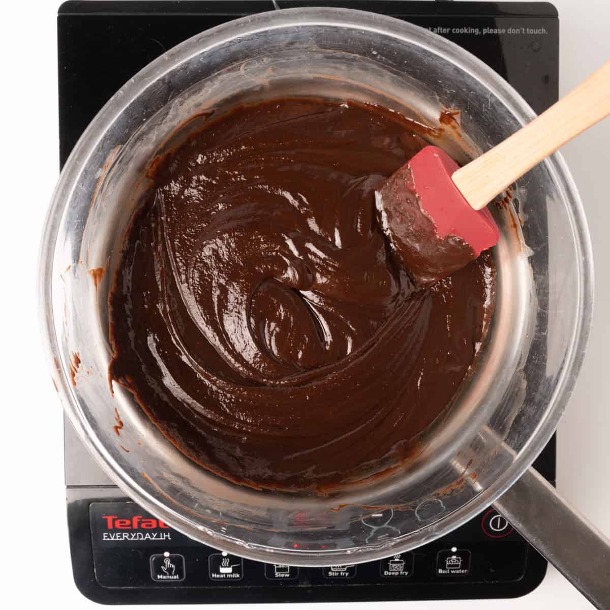 A bowl of thick chocolate mixture, stirred over a pan of hot water on an electric stove.
