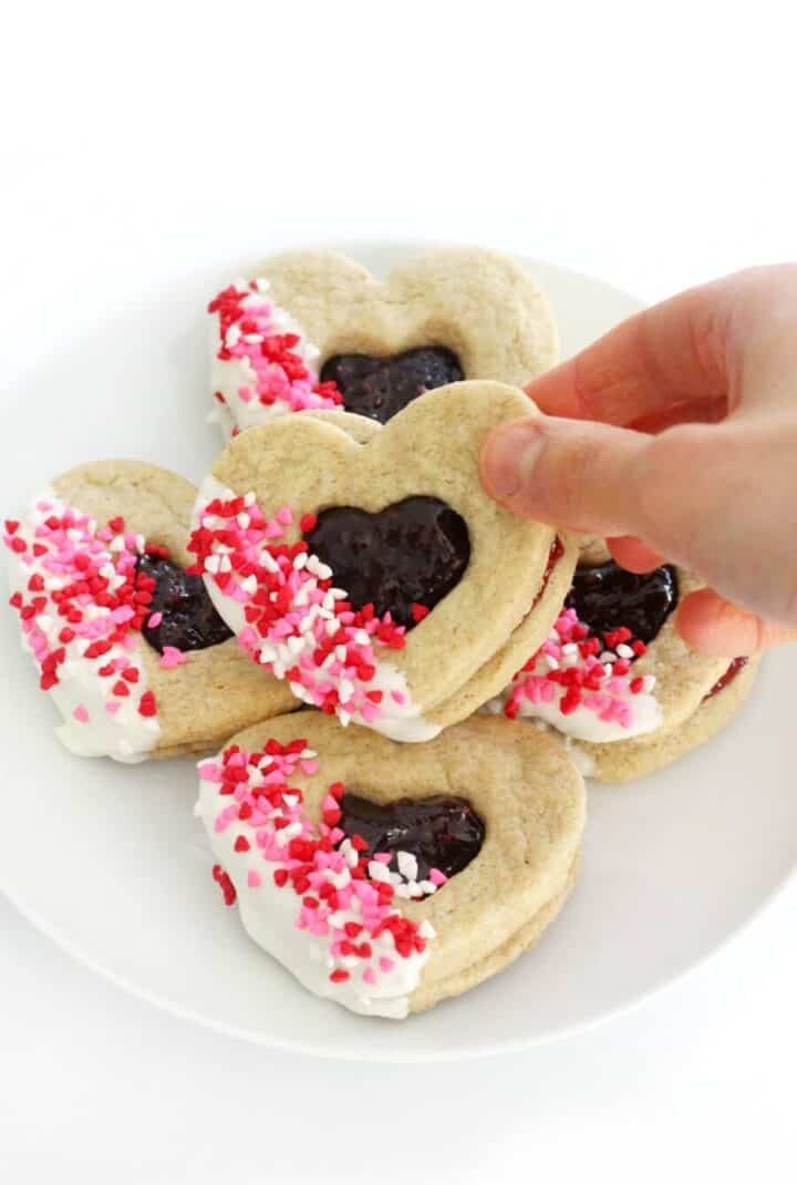 A hand lifts a heart shaped sandwich cookie from a plate.
