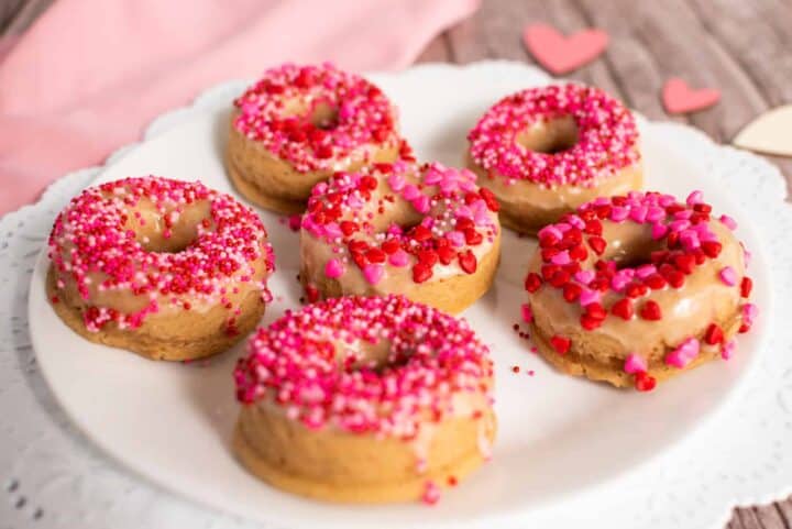 Six donuts on a plate covered in pink and heart shaped sprinkles.