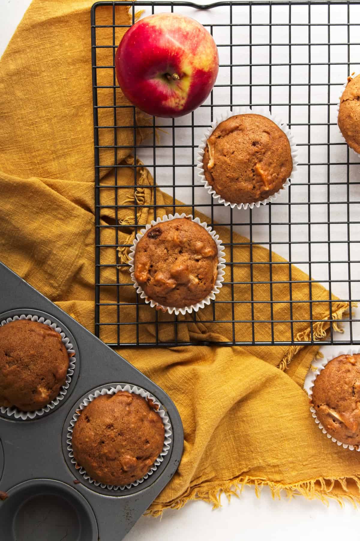 Muffins on a cooling rack next to a muffin tray with a yellow cloth.