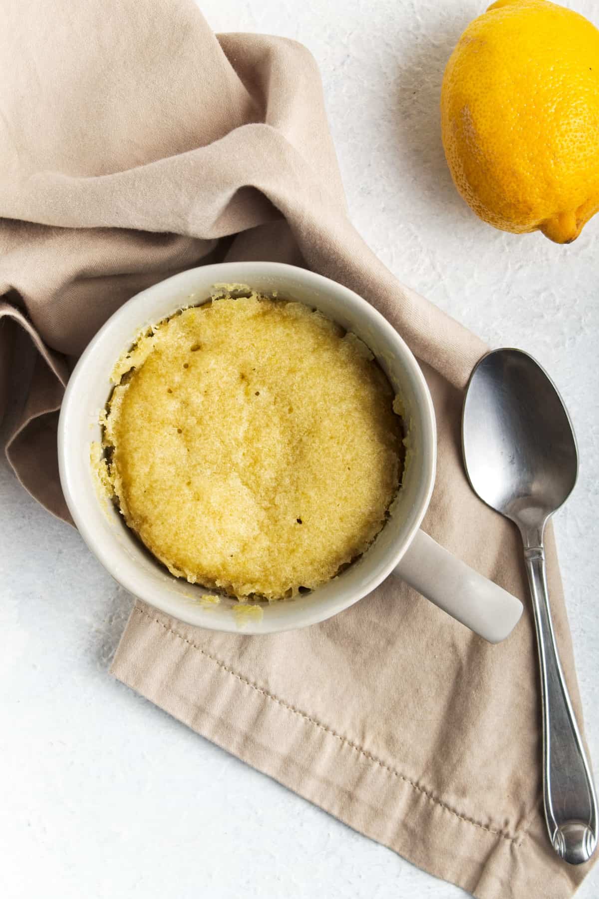 A cake in a mug next to a spoon, a lemon and a napkin.
