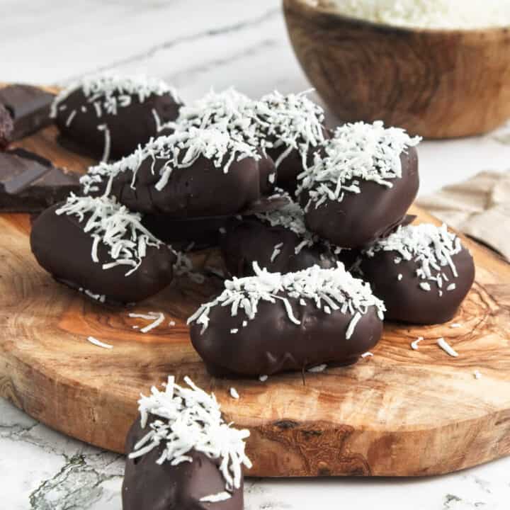 Chocolate covered dates topped with coconut on a wooden board.