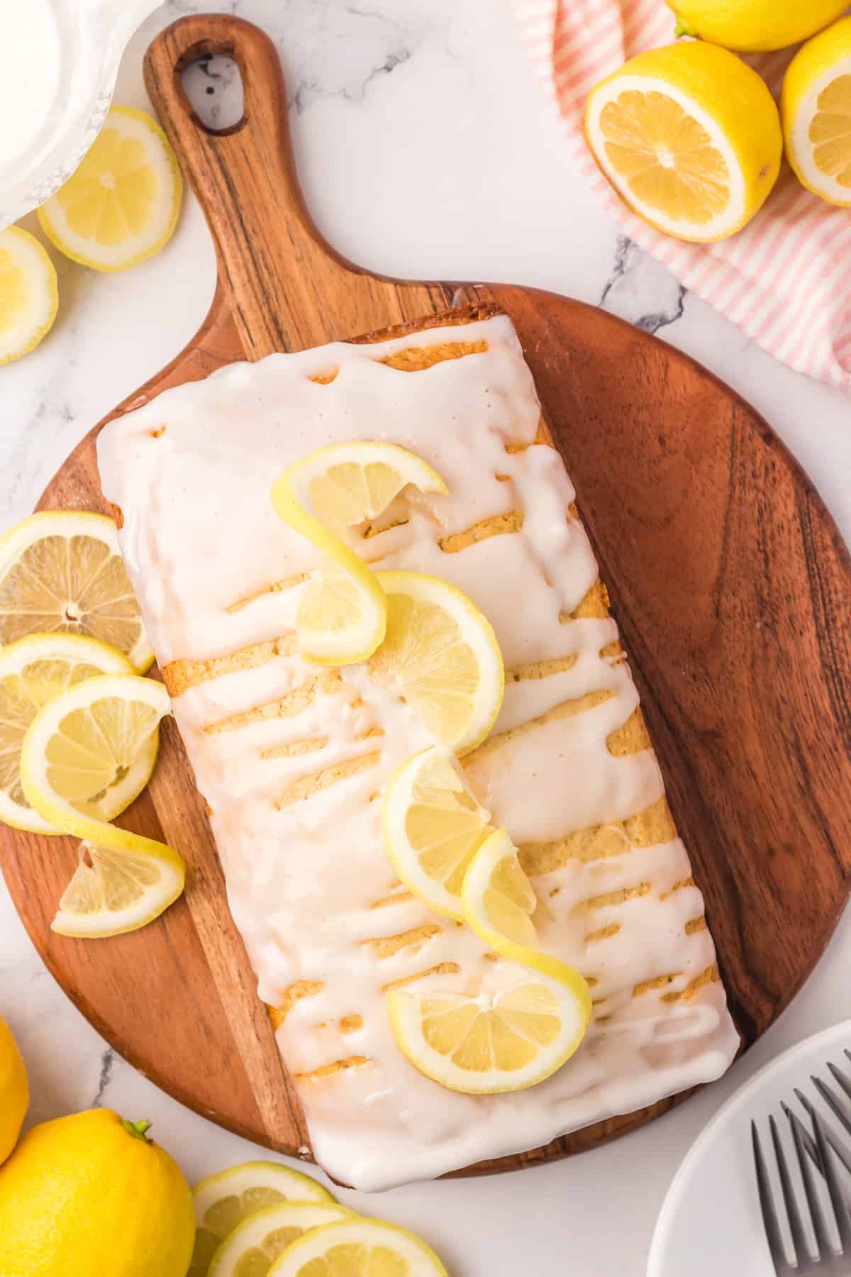 Vegan lemon loaf topped with glaze and lemon slices on a round wooden board.
