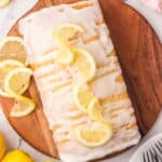 Vegan lemon loaf topped with glaze and lemon slices on a round wooden board.