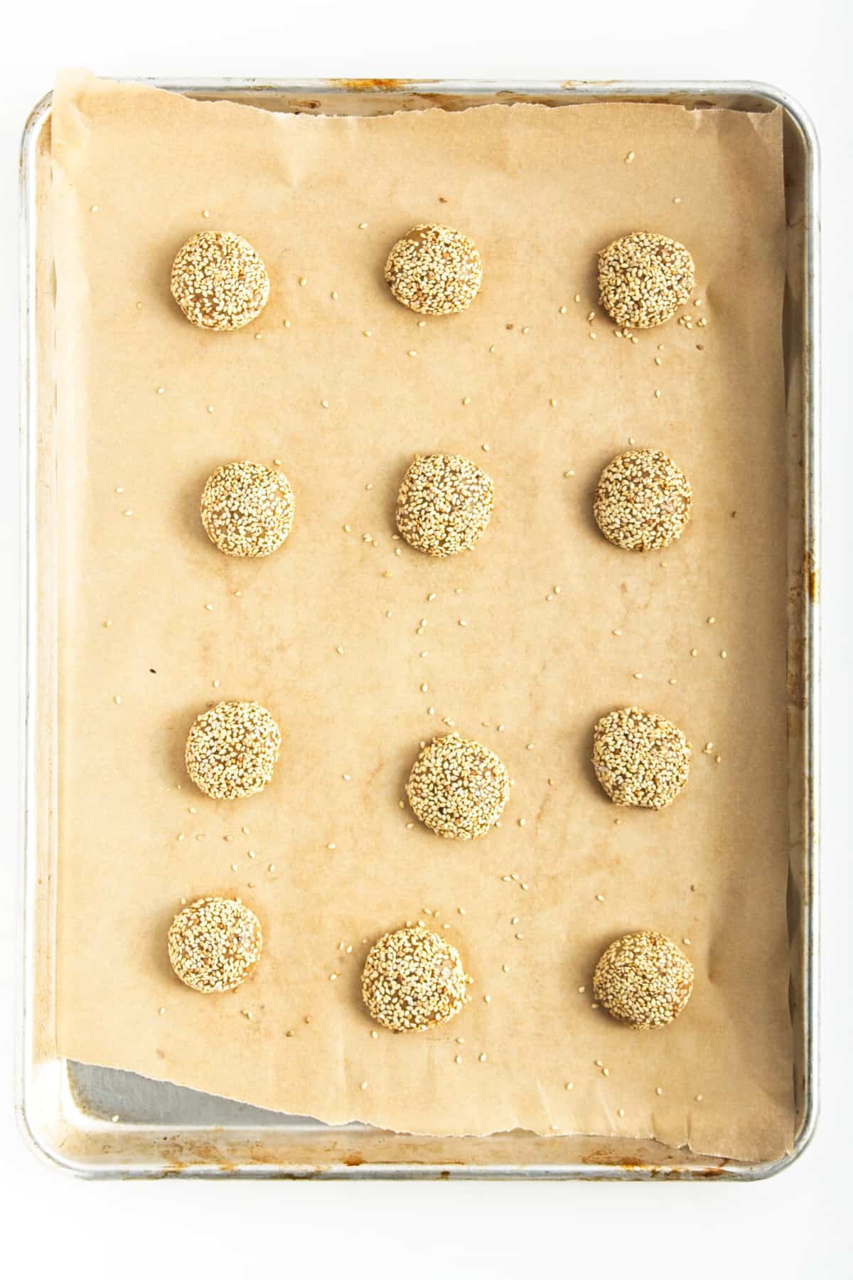 Cookies rolled in sesame seeds on a lined baking sheet.